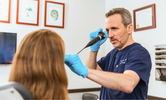 A doctor in navy scrubs, wearing blue gloves, holds a medical tool, attentively speaking to a seated patient in an office. Certificates are on the wall.