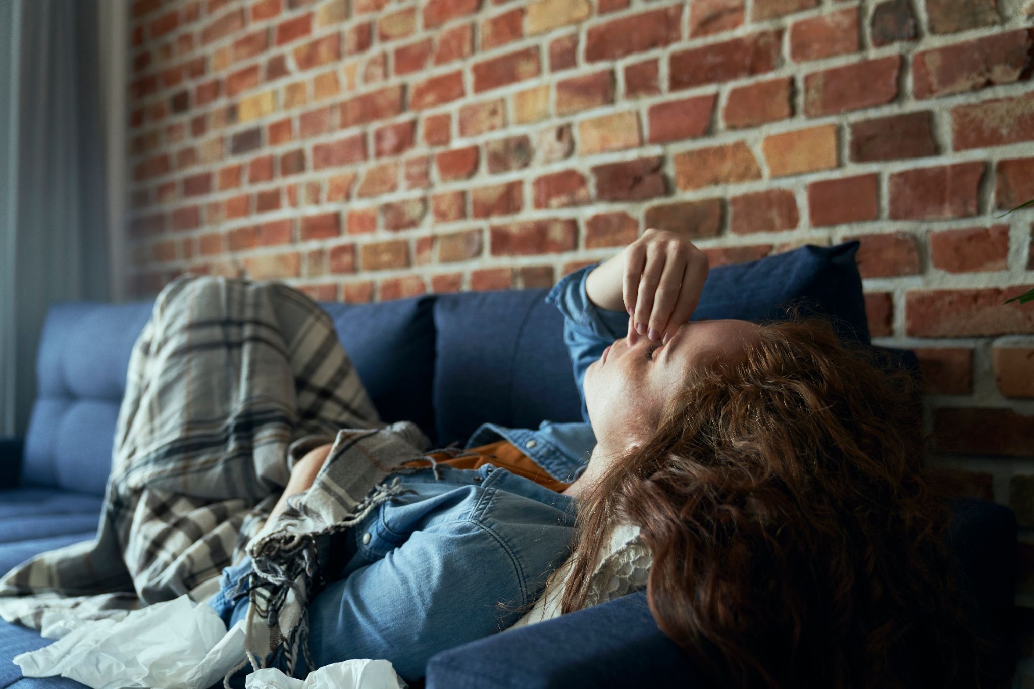 A tired person lies curled on a blue couch, covering their eyes, with long brown hair and a plaid blanket against a brick wall.