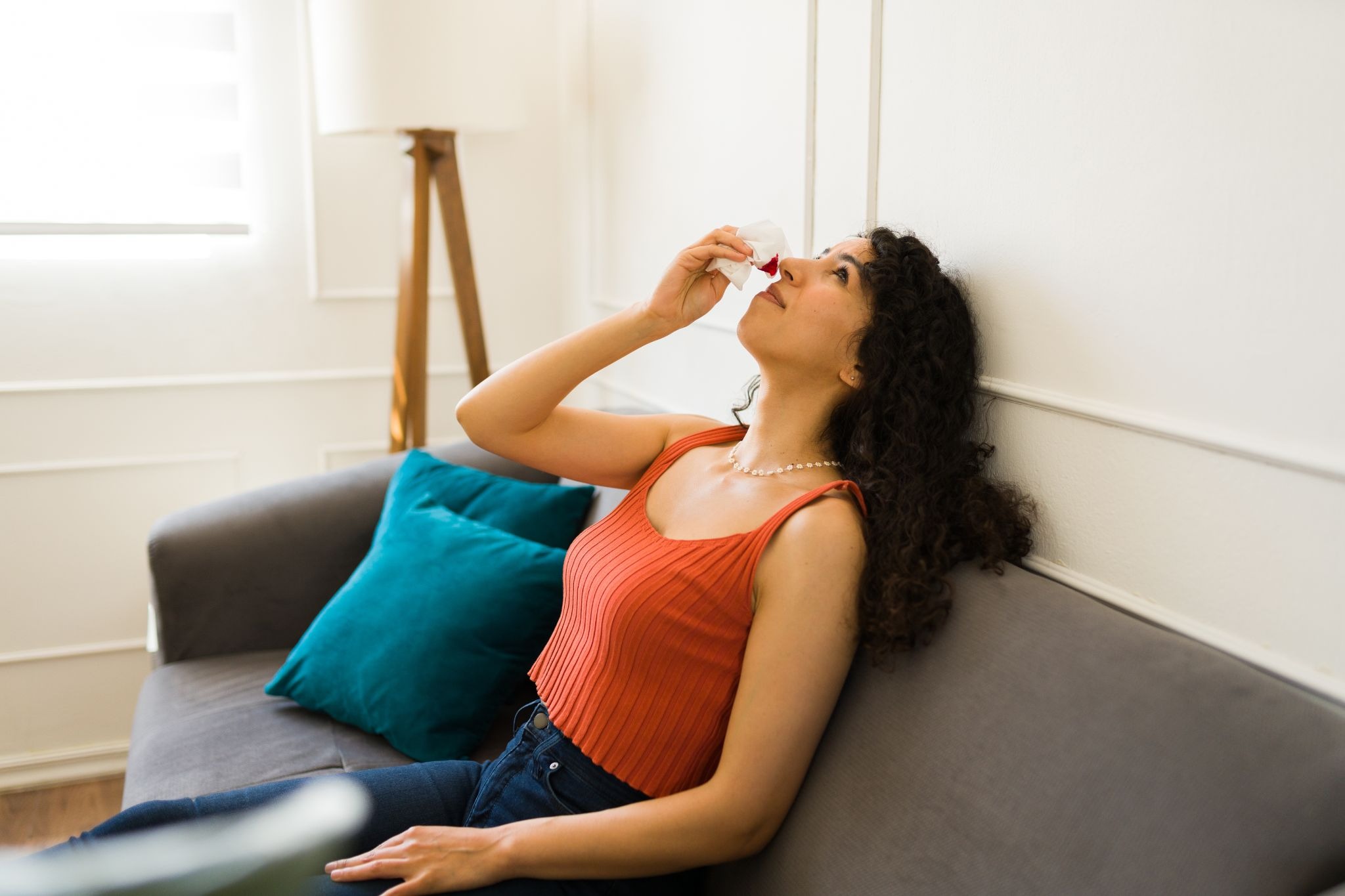 A woman on a couch holding a bloody tissue to her nose, tilting her head back