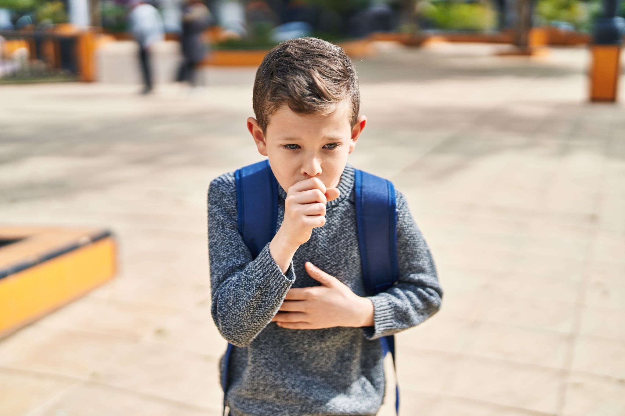 Young boy in a gray sweater and blue backpack covers his mouth while coughing in an outdoor schoolyard, conveying discomfort and concern.