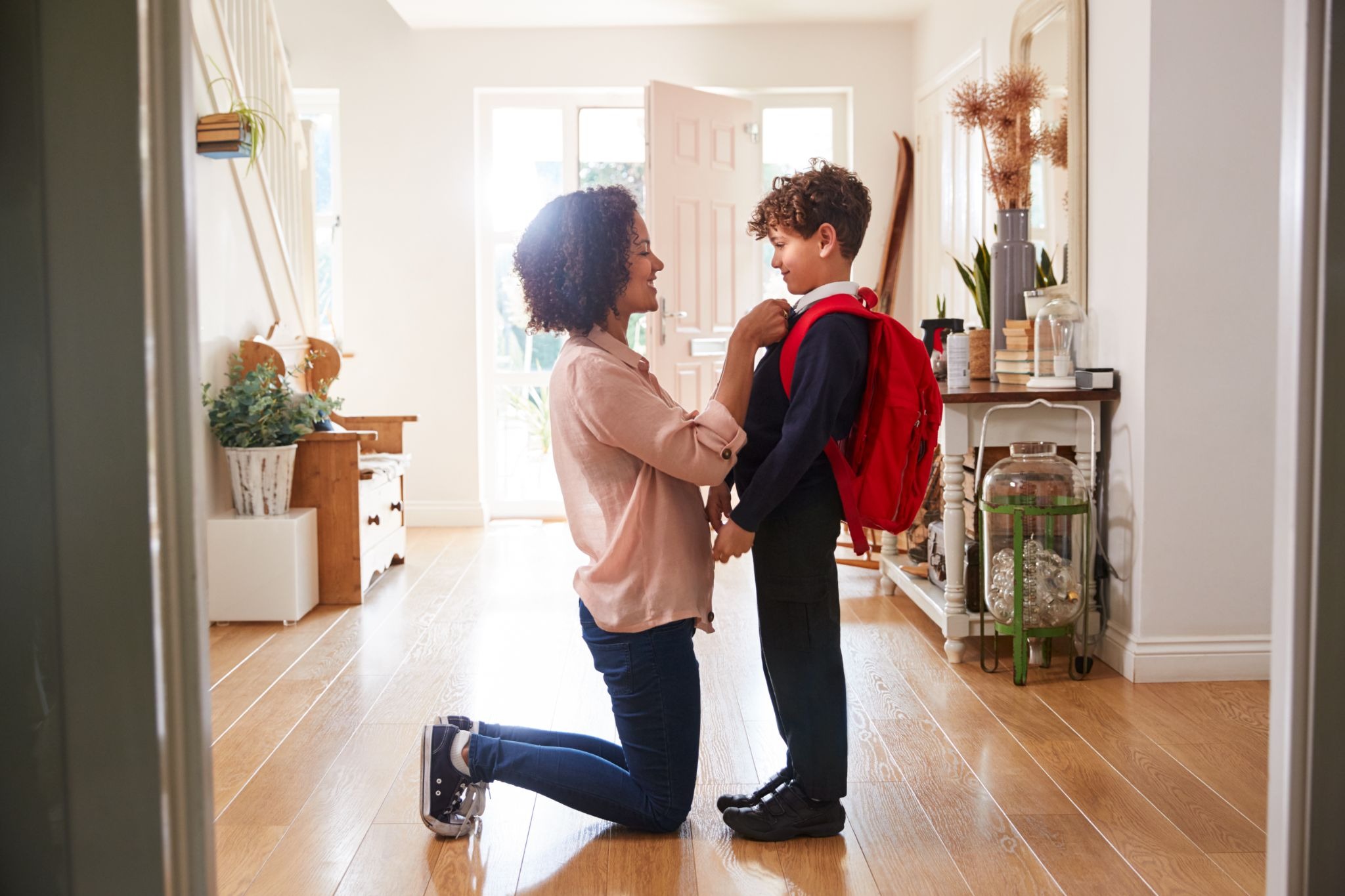 A mother kneels to hug her smiling school-age son wearing a red backpack in a bright, welcoming home entryway.