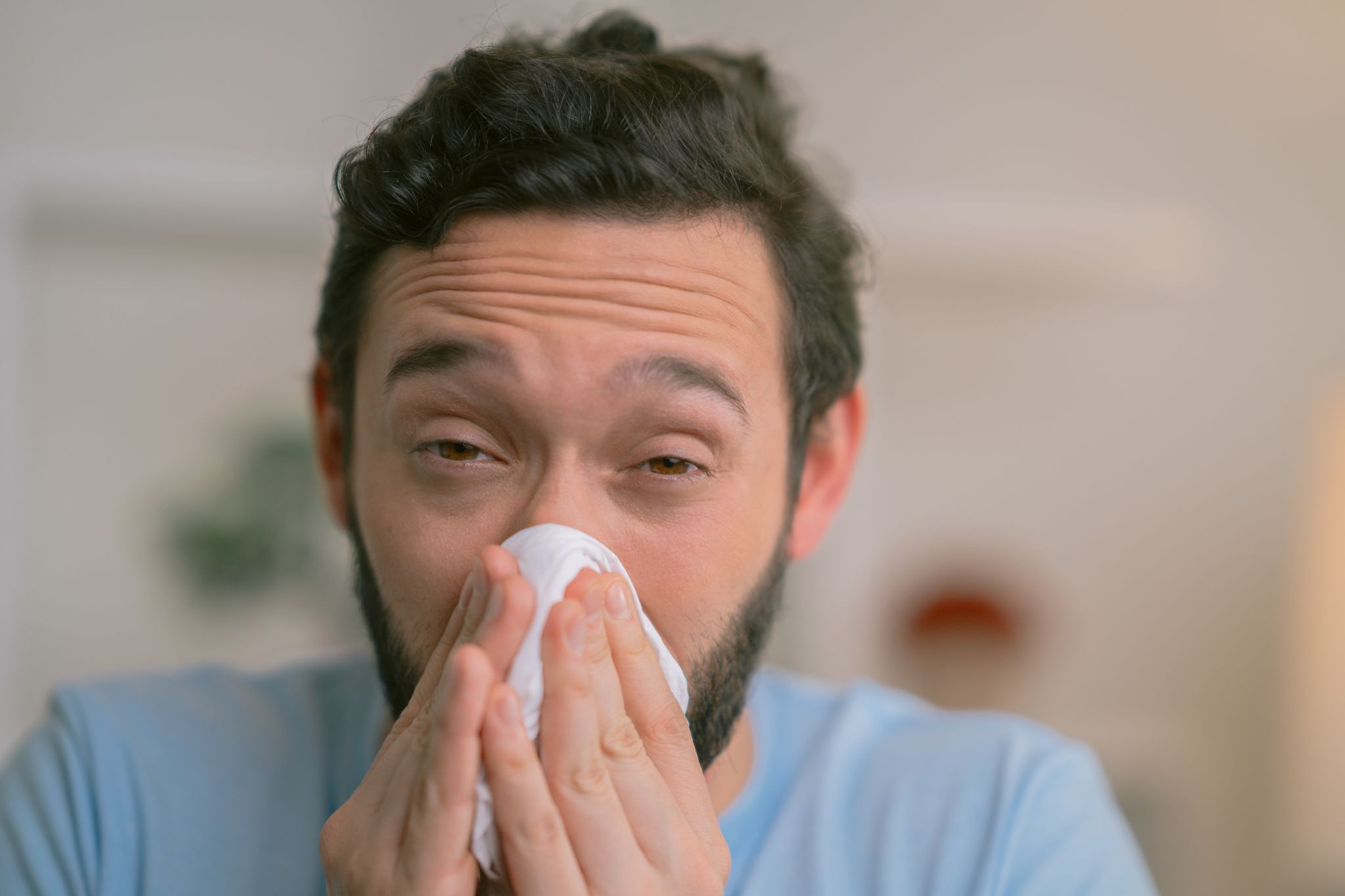 Man with dark hair and a beard sneezes into a white tissue at home, eyes narrowed and face flushed, conveying illness and discomfort.