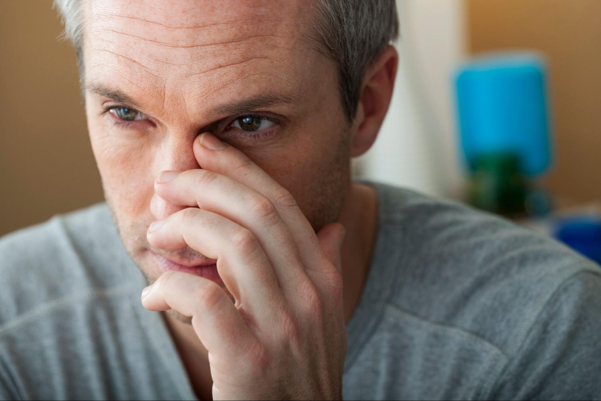 Close-up of a tired, pensive man rubbing his eye and nose, with a blurred home interior behind him, conveying stress or sadness.